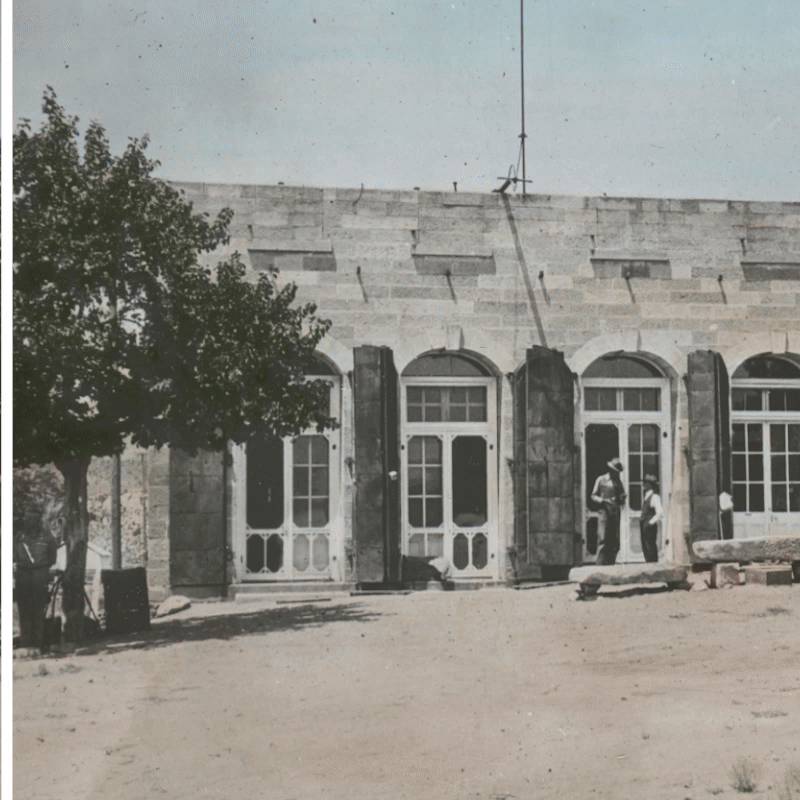 Two people stand outside a stone building with arched doors and windows, next to a tree.