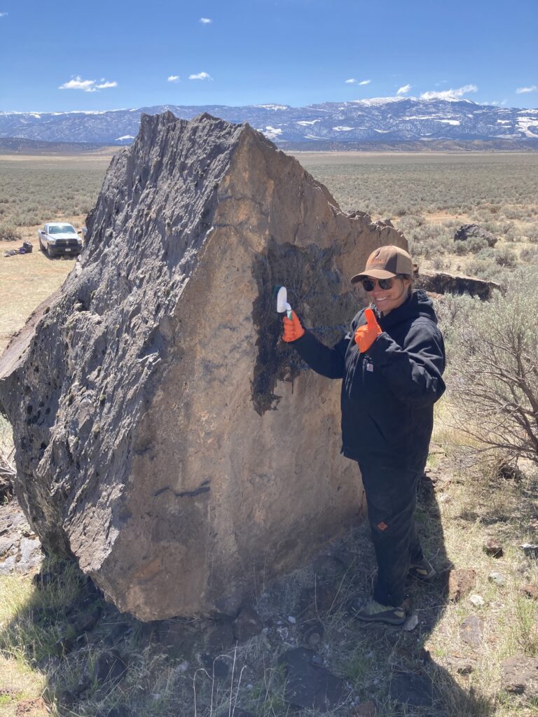 Person in outdoor gear studies a large rock with tools in a grassy, mountainous landscape.