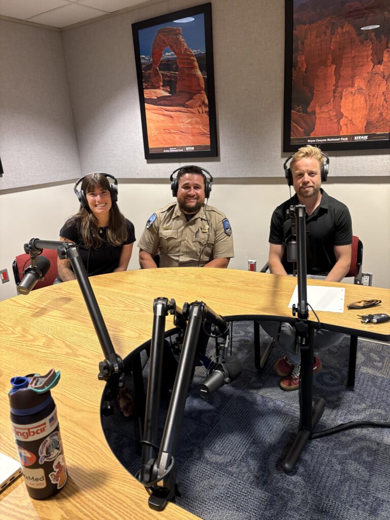 Three people with headsets sit at a podcast table with microphones in a studio setting.