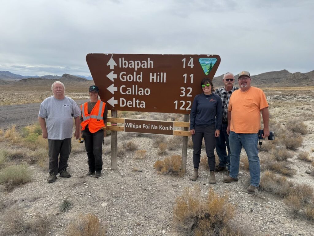 Five people stand by a road sign listing distances to Ibapah, Gold Hill, Callao, and Delta.