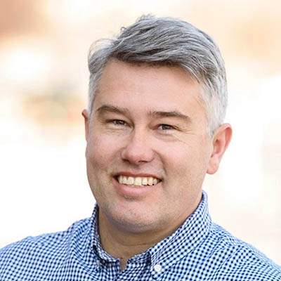 Smiling man with short gray hair wearing a blue checkered shirt, outdoors.