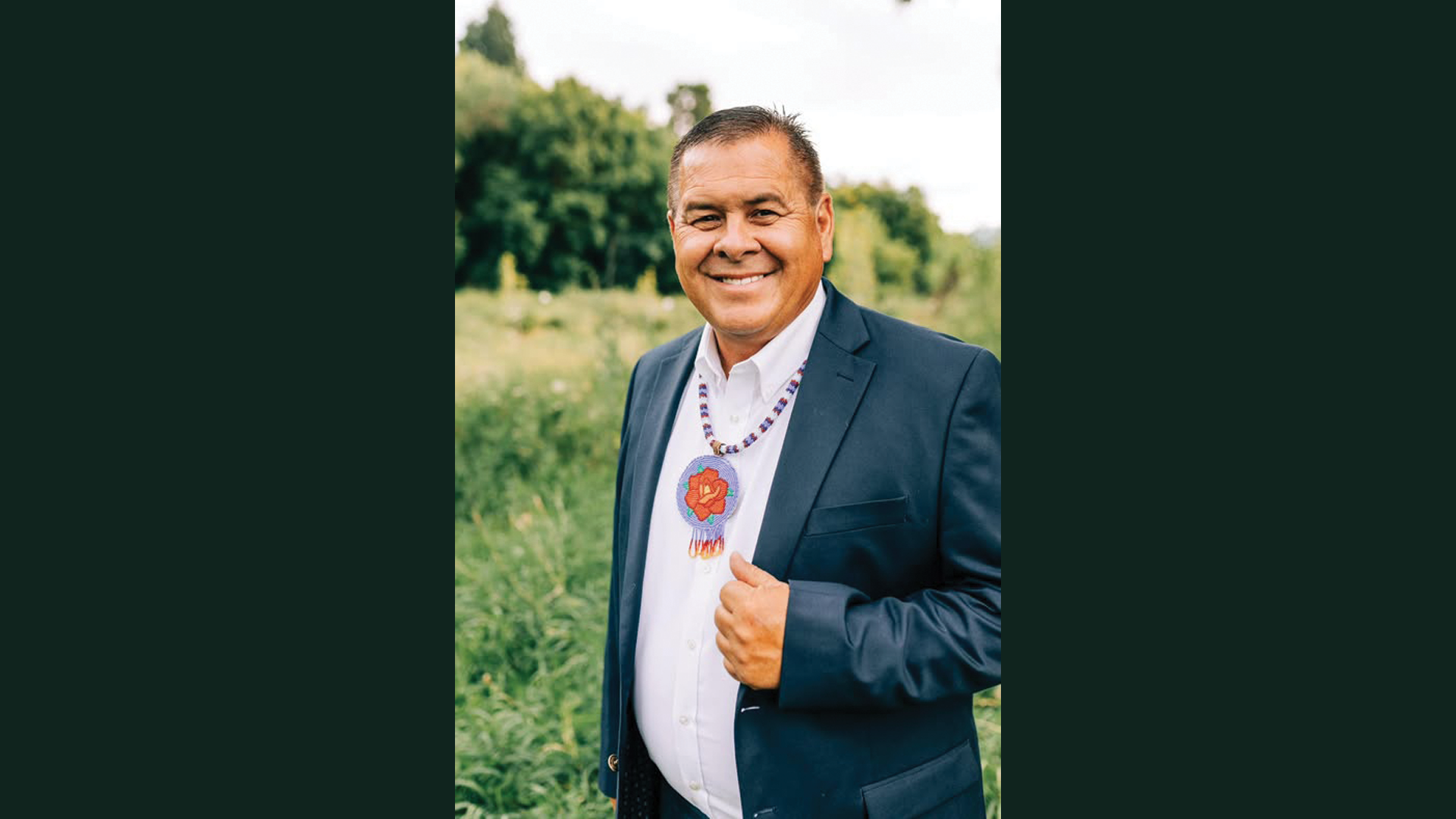 Smiling man in a suit with a beaded necklace, standing outdoors in a green field.