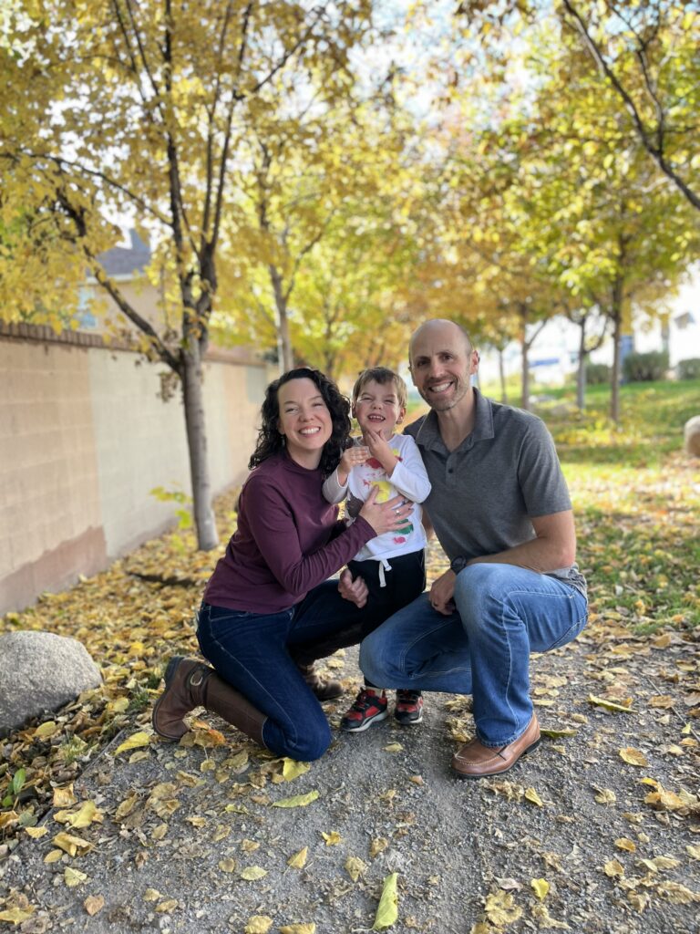 A smiling family of three poses together on a leafy path under autumn trees.