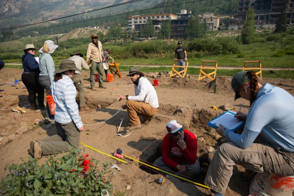 People work at an outdoor archaeological dig site with tools, buckets, and measuring tape visible.