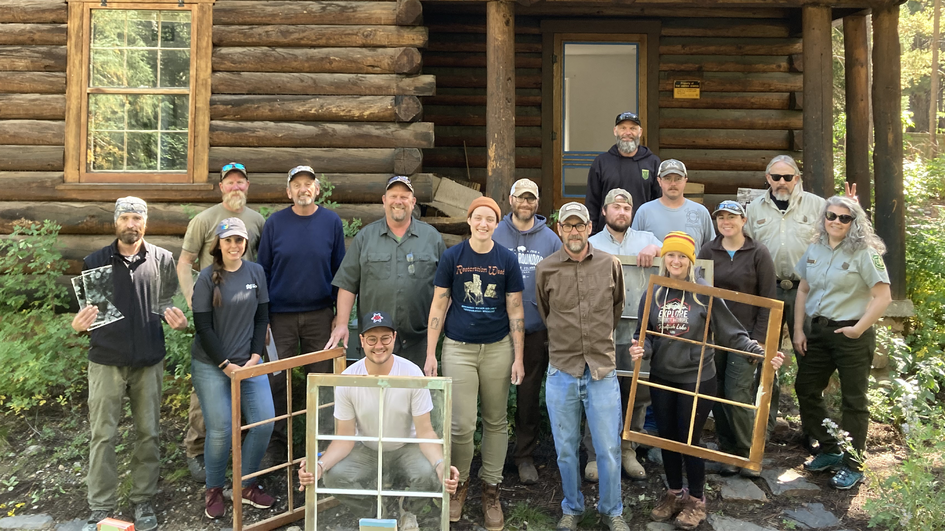 Group of people posing in front of a log cabin, some holding window frames, outdoors.