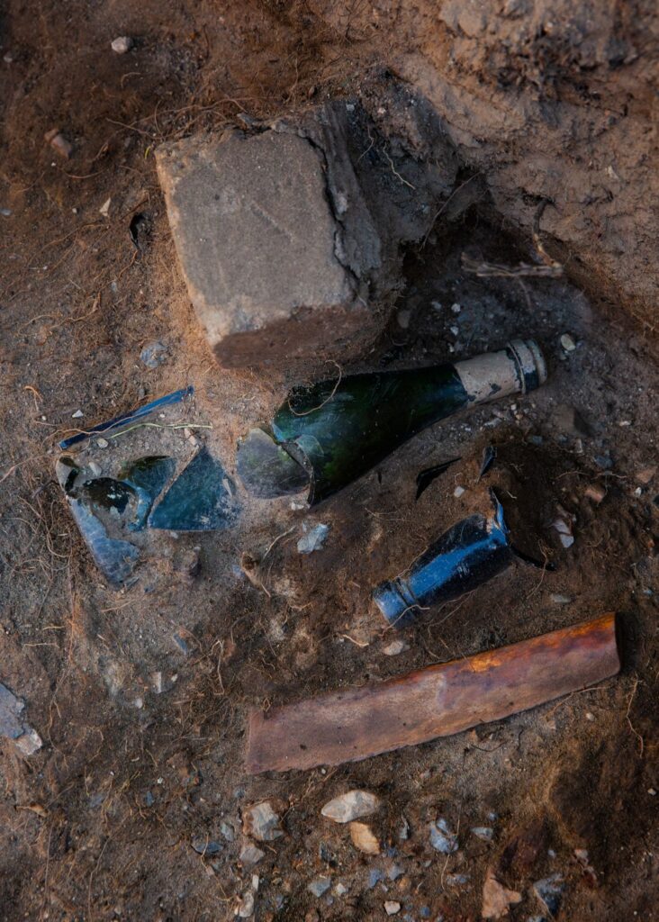 Broken glass bottles, a rusted metal piece, and a brick lie on dirt and rocks.