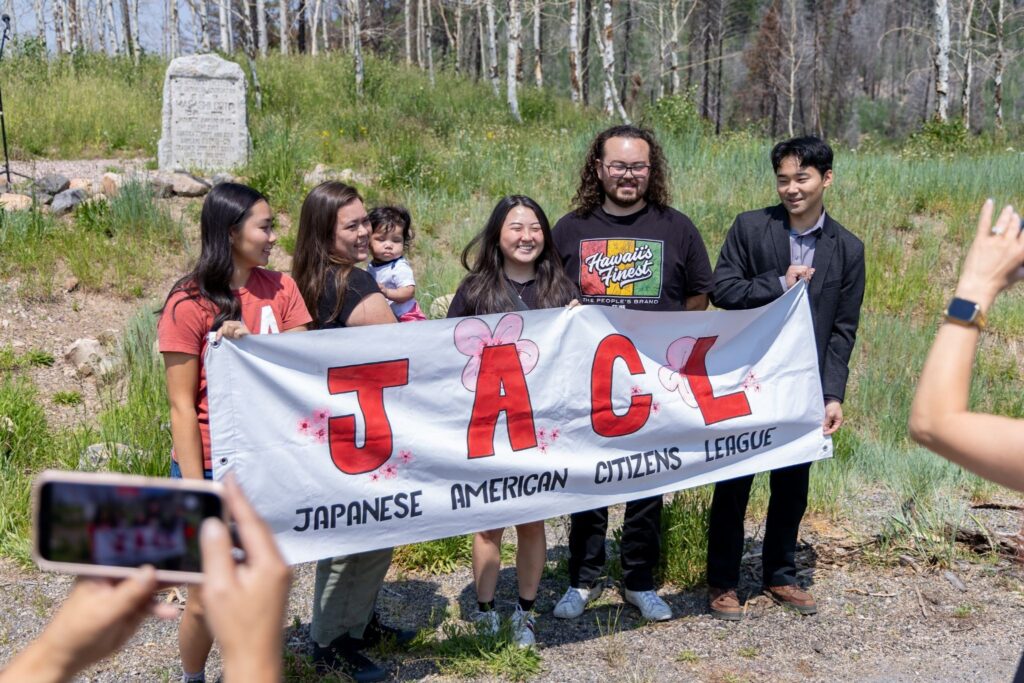 A group holds a Japanese American Citizens League banner outdoors while someone takes their photo.