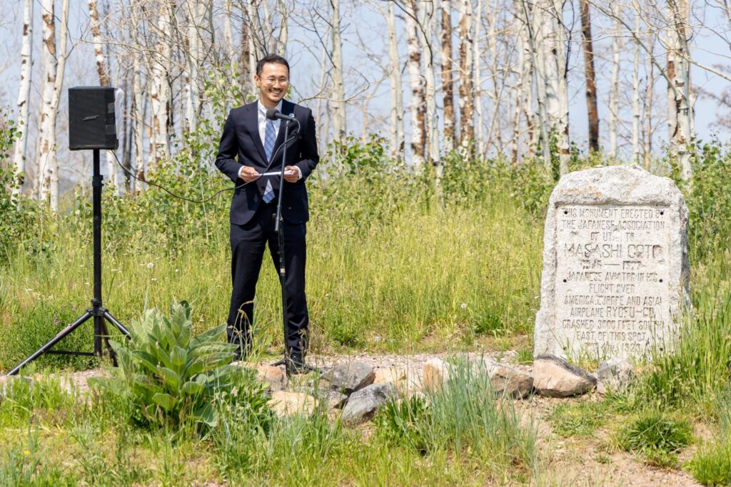 A man in a suit speaks at a podium near a stone memorial in a grassy area with trees.